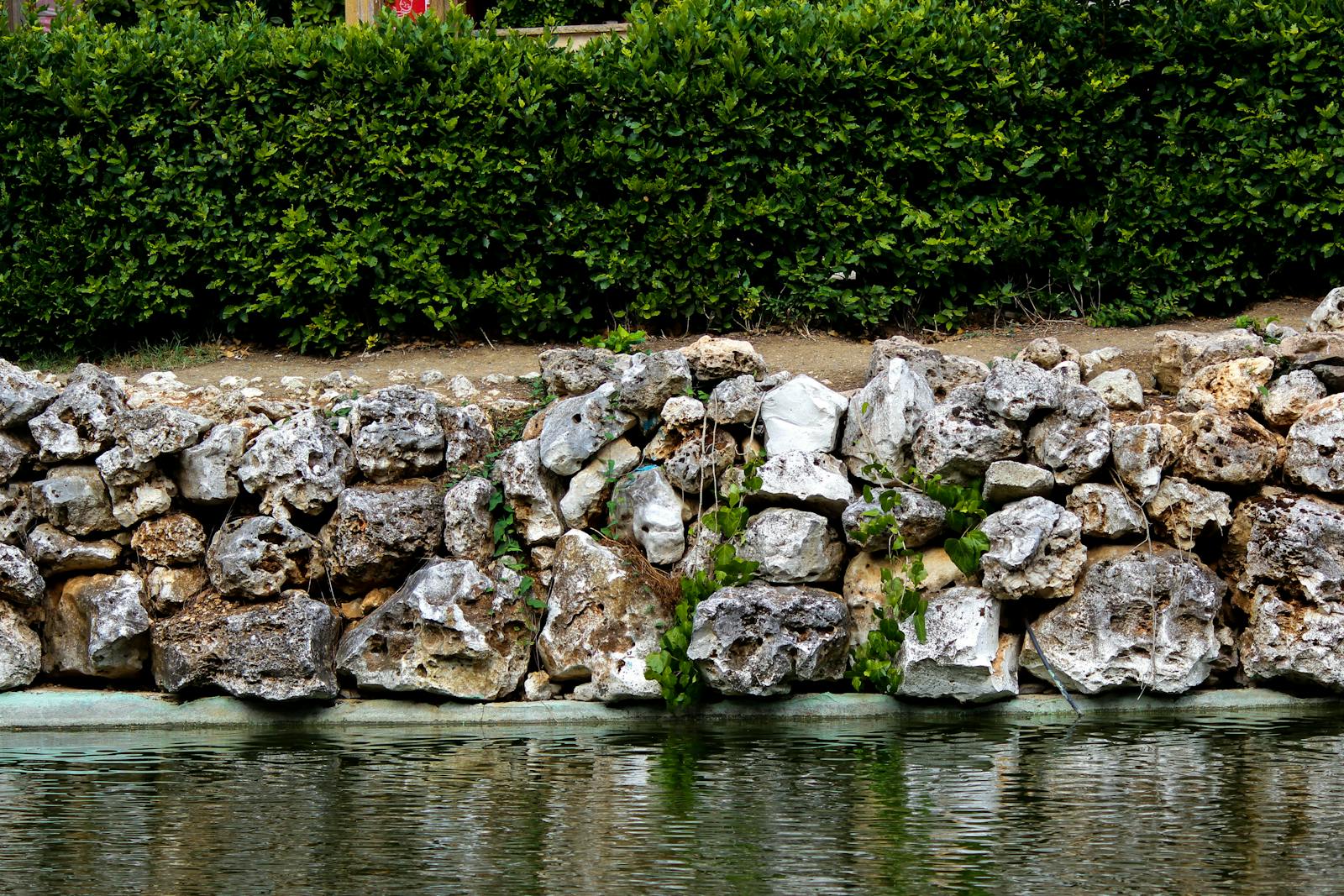 Mortared-stone retaining wall above a small pond, with clipped hedges behind.